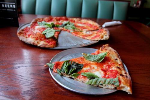 Two pizzas on metal trays, one with slices missing, topped with tomato sauce and fresh basil, on a wooden table.
