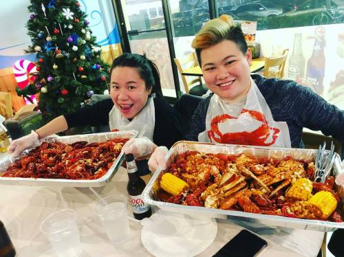 Two women smiling and holding large trays of seafood, including crabs and corn, in a festive restaurant setting.