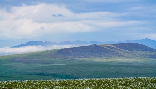 A serene landscape featuring rolling green hills under a cloudy blue sky, with distant mountains in the background.