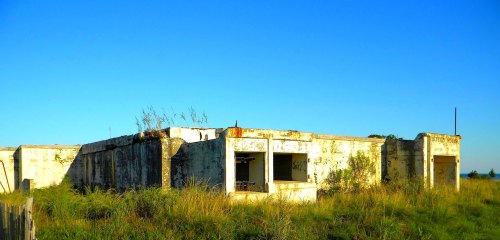 Abandoned building with crumbling walls, overgrown grass, and a clear blue sky in the background.