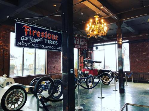 Vintage cars displayed in a rustic setting with banners for Firestone tires and Willard service station.
