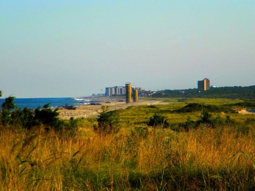 A coastal landscape featuring sandy shores, distant buildings, and grassy dunes under a clear sky.