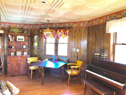 Cozy room with wooden walls, a table, yellow chairs, a piano, and floral curtains by the windows.