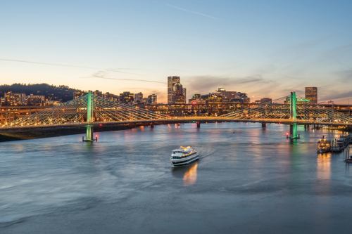 A boat sails under a brightly lit bridge with a city skyline at dusk, reflecting on the water.