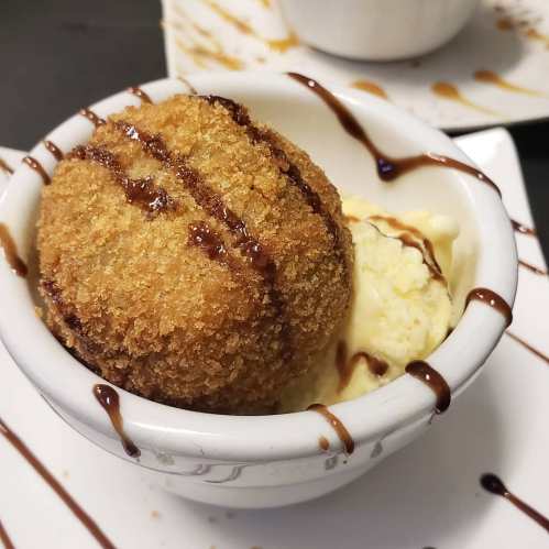 A bowl with a fried dessert ball on top of vanilla ice cream, drizzled with chocolate sauce.