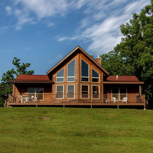 A large wooden house with a sloped roof and large windows, surrounded by green grass and trees under a blue sky.