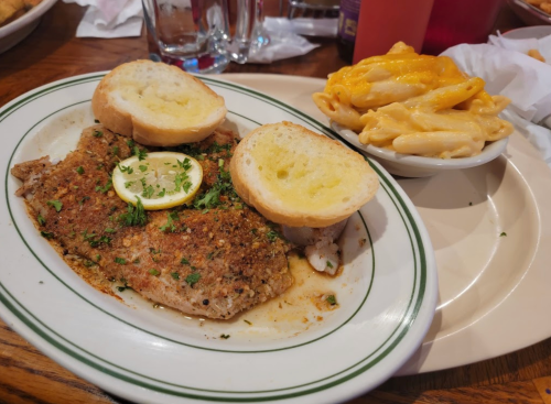 A plate featuring seasoned fish with lemon and two slices of garlic bread, alongside a bowl of creamy macaroni and cheese.