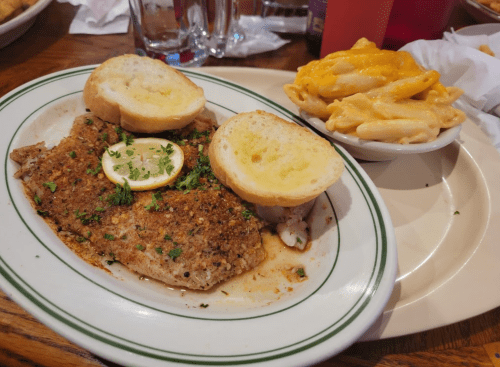 A plate featuring seasoned fish with lemon and two slices of garlic bread, alongside a bowl of creamy macaroni and cheese.