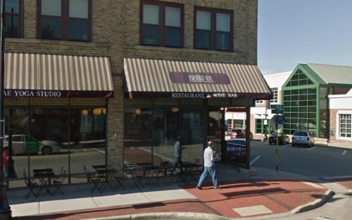 A street view of a restaurant and wine bar with outdoor seating, next to a yoga studio and modern buildings.