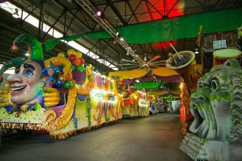 Colorful parade floats decorated with masks, balloons, and festive designs inside a large warehouse.