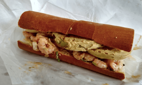 A shrimp sandwich on a crusty roll, filled with shrimp, lettuce, and a creamy spread, wrapped in white paper.