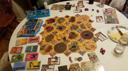A tabletop game setup featuring a hexagonal board, various game pieces, cards, and dice on a dining table.