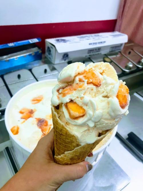 A hand holding a large ice cream cone topped with peach-flavored ice cream, with a tub of ice cream in the background.