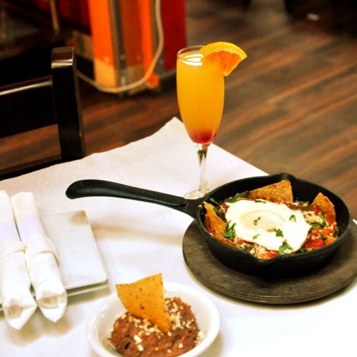 A skillet of nachos topped with eggs and herbs, a bowl of refried beans, and a colorful drink with an orange slice.
