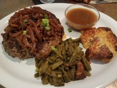 A plate featuring a juicy hamburger topped with caramelized onions, green beans, cornbread, and a side of gravy.