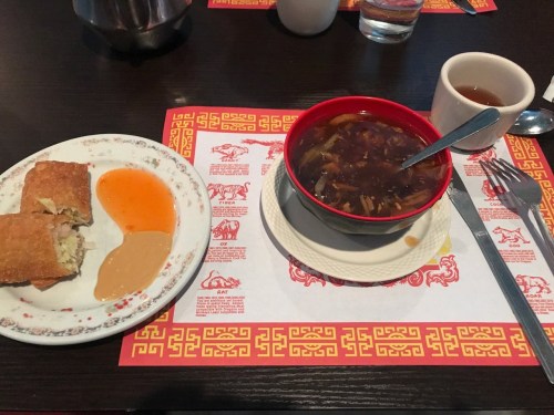A plate with two spring rolls and sauces, alongside a bowl of soup and a cup of tea on a patterned table mat.