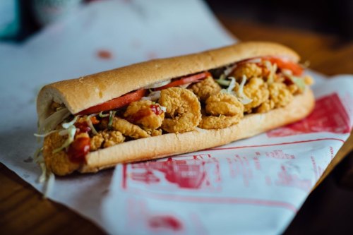 A long sandwich filled with fried shrimp, lettuce, tomatoes, and ketchup, served on a paper wrapper.