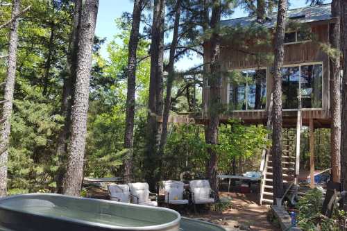 A treehouse nestled among tall trees, with outdoor seating and a bathtub in the foreground, surrounded by greenery.