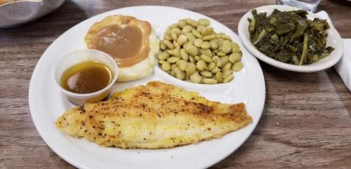 A plate of fried fish, collard greens, lima beans, a biscuit with gravy, and a small cup of sauce.