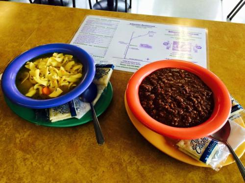 Two colorful bowls on a table: one with noodle soup and the other with chili, accompanied by packets of crackers.