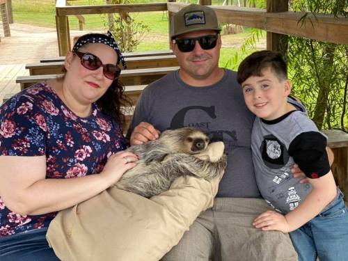 A family of three sits together, holding a sloth on a blanket, smiling in a natural outdoor setting.