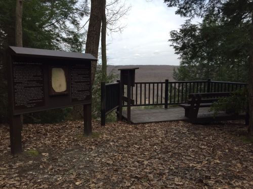 Viewing platform in a wooded area, featuring informational signs and a bench overlooking a valley.