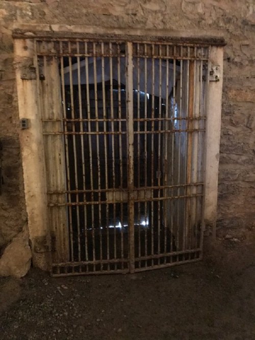 Rusty barred gate leading to a dark, stone-walled passageway, with faint light visible through the bars.