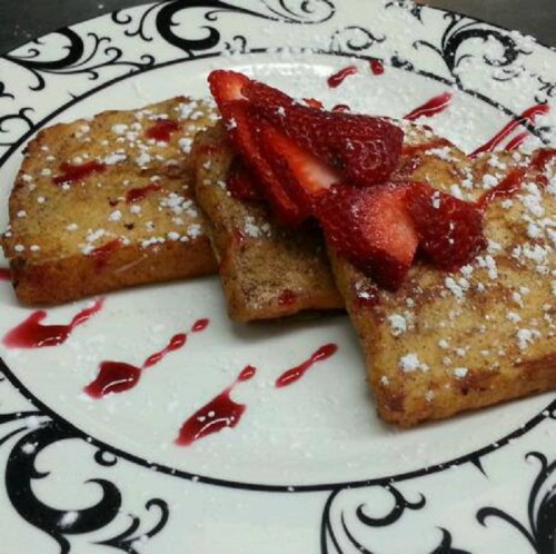 Three slices of French toast topped with strawberries and powdered sugar, drizzled with berry sauce on a decorative plate.