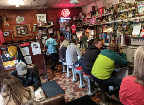 A busy diner with patrons sitting at the counter and tables, surrounded by vintage decor and a neon sign.