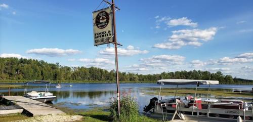 A lakeside scene featuring a sign for food and spirits, with boats docked and lush greenery under a blue sky.