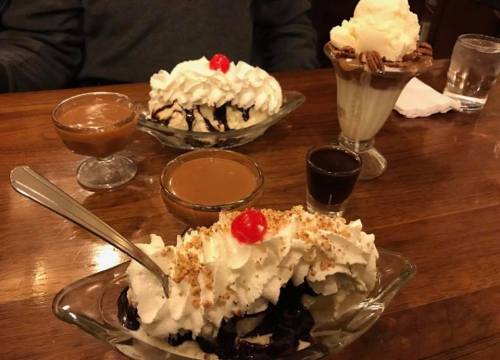 Three dessert bowls on a wooden table: a banana split, a sundae, and a small cup of chocolate sauce.