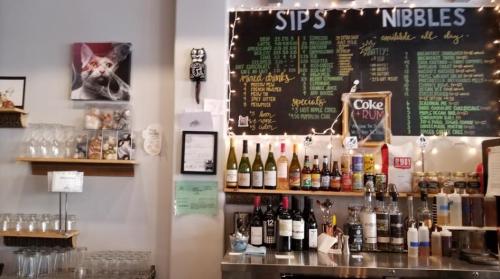 A cozy bar with a chalkboard menu, shelves of drinks, and a cat painting on the wall, adorned with string lights.