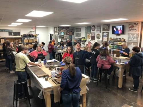 A lively workshop with people crafting wooden signs, smiling, and engaging in creative activities at various tables.