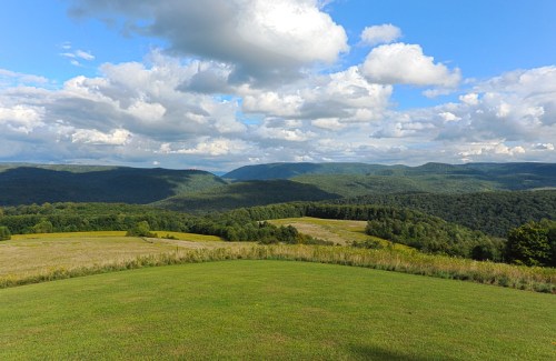 A panoramic view of rolling green hills and mountains under a blue sky with fluffy white clouds.