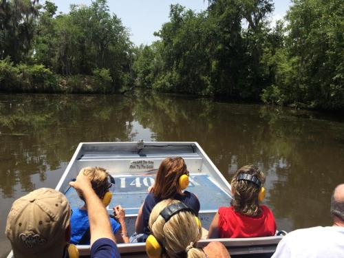 A group of people wearing ear protection on a boat, navigating a calm, tree-lined waterway on a sunny day.