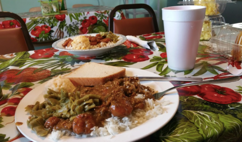 A plate of rice with green beans, meat, and a slice of bread, with a cup and another plate of food in the background.