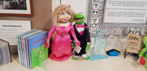 A Kermit the Frog and Miss Piggy doll in formal attire, surrounded by children's books and awards on a display table.