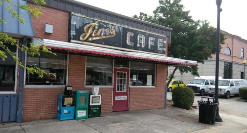 A vintage cafe with a neon sign, featuring a red door and newspaper stands outside, set in a small town.