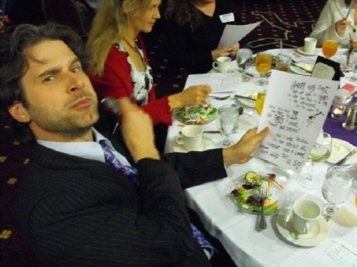 A man in a suit holds a paper while sitting at a dining table with food and drinks, surrounded by other guests.