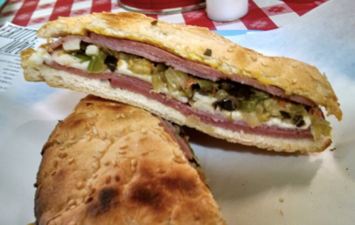 A close-up of a sandwich filled with layers of meats, pickles, and mustard, resting on a paper-lined plate.