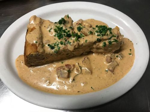 A slice of savory dish topped with creamy sauce and herbs, served on a white plate with a rich, flavorful sauce underneath.