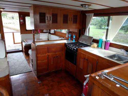 Interior of a wooden boat kitchen, featuring a stove, sink, cabinets, and a cozy seating area with large windows.