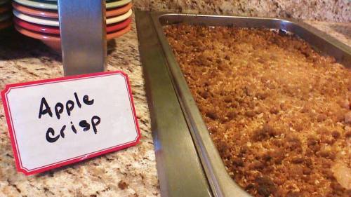 A tray of apple crisp with a handwritten sign next to it on a countertop.