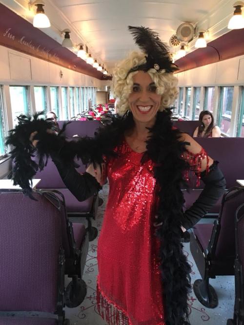 A person in a red sequined dress and black feather boa smiles in a vintage train car filled with passengers.