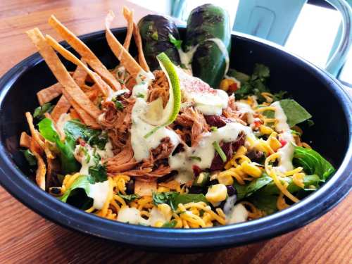 A colorful salad bowl with pulled chicken, tortilla strips, cheese, greens, jalapeños, and a drizzle of dressing.