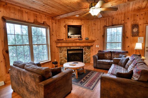 Cozy living room with wooden walls, a stone fireplace, two sofas, a coffee table, and large windows.