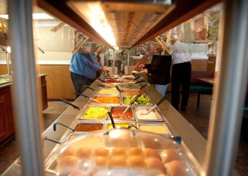 A buffet with various food options, including salads, sauces, and rolls, with diners serving themselves in the background.