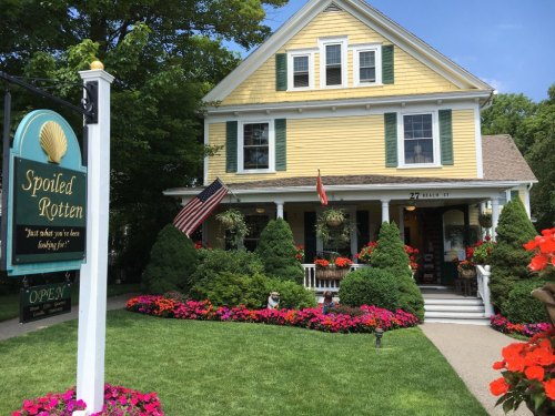 A charming yellow house with green shutters, surrounded by colorful flowers and a sign that reads "Spoiled Rotten."