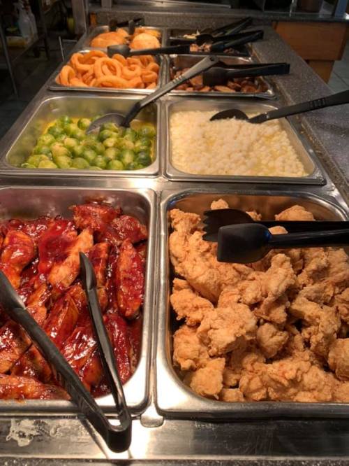 A buffet spread featuring fried chicken, BBQ ribs, Brussels sprouts, onion rings, and rice in metal trays.