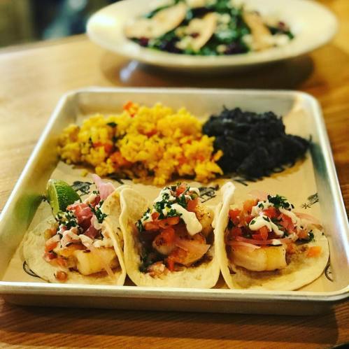 Three tacos topped with shrimp, pico de gallo, and sour cream, served with yellow rice and black beans on a tray.